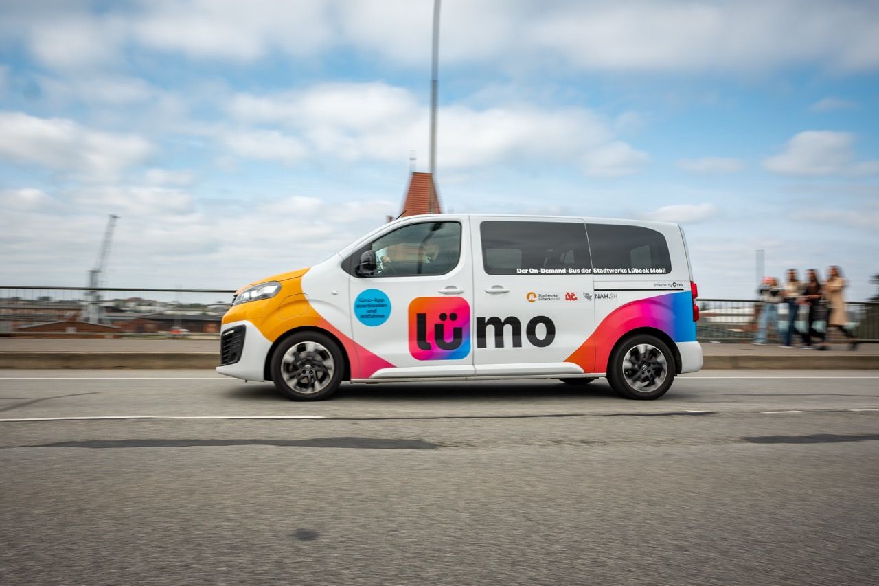 A colorful van with "Lümo" branding drives on a road, featuring vibrant graphics and logos, with a cityscape and cloudy sky in the background.