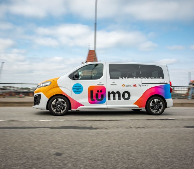 A colorful van with "Lümo" branding drives on a road, featuring vibrant graphics and logos, with a cityscape and cloudy sky in the background.