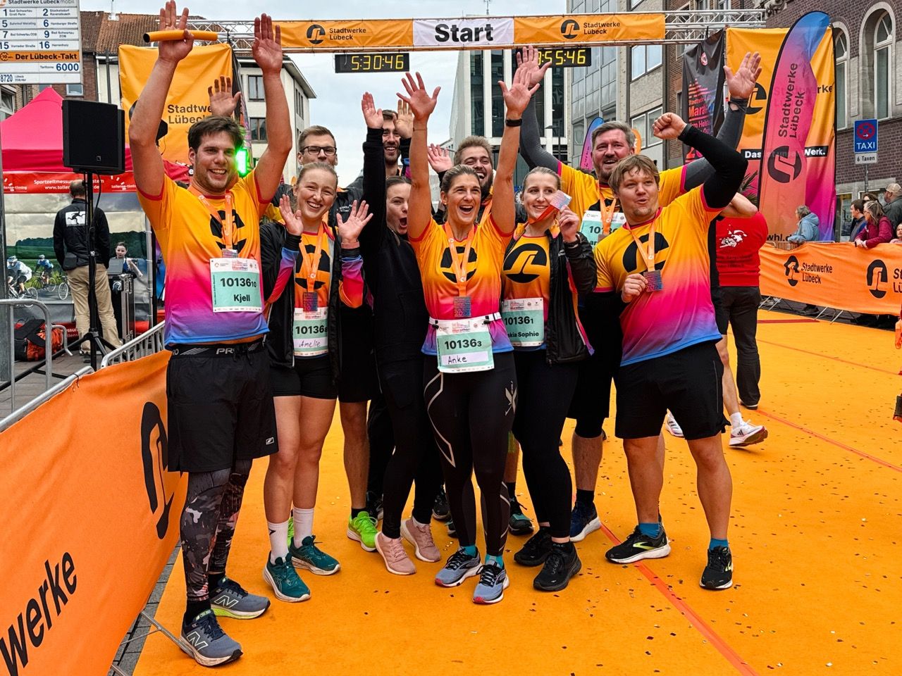 A group of runners in colorful shirts celebrate at a race finish line, raising their arms in excitement.