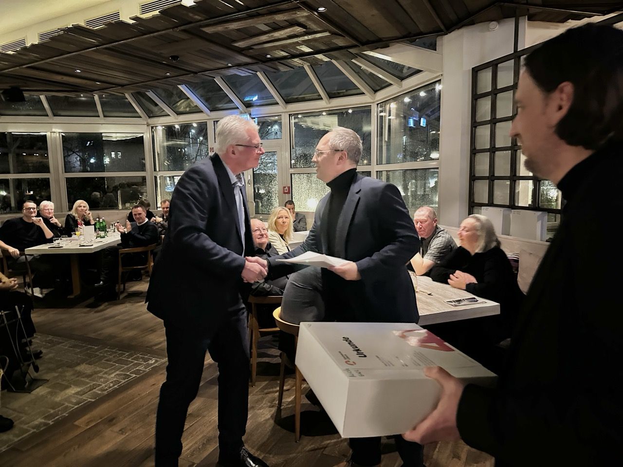 Two men shake hands in a restaurant, surrounded by seated people. One man holds a certificate, while another holds a box.