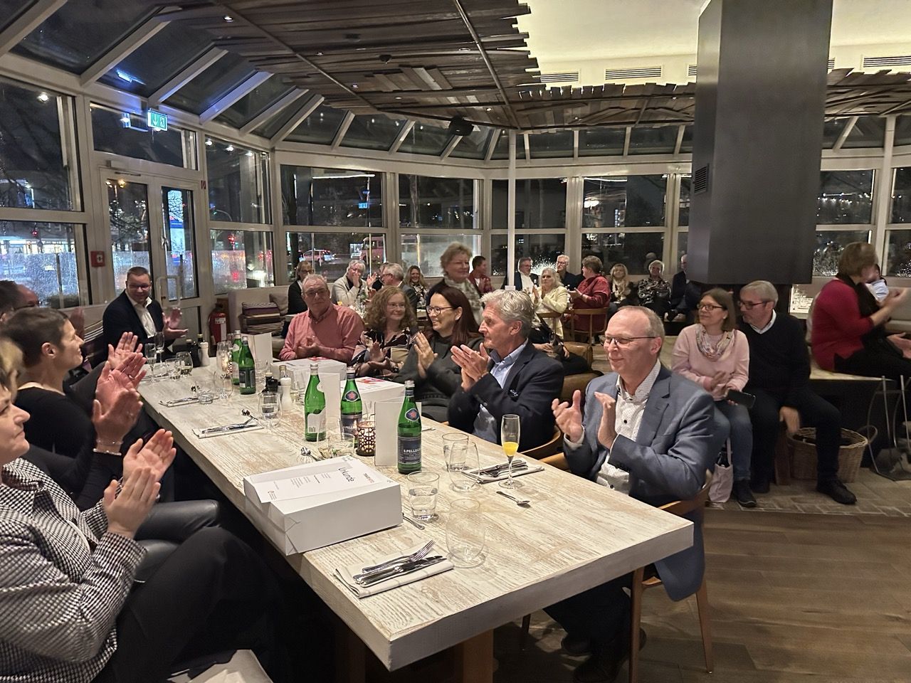 People seated in a restaurant, clapping and smiling. Tables have drinks and food. Large windows show a nighttime city view.