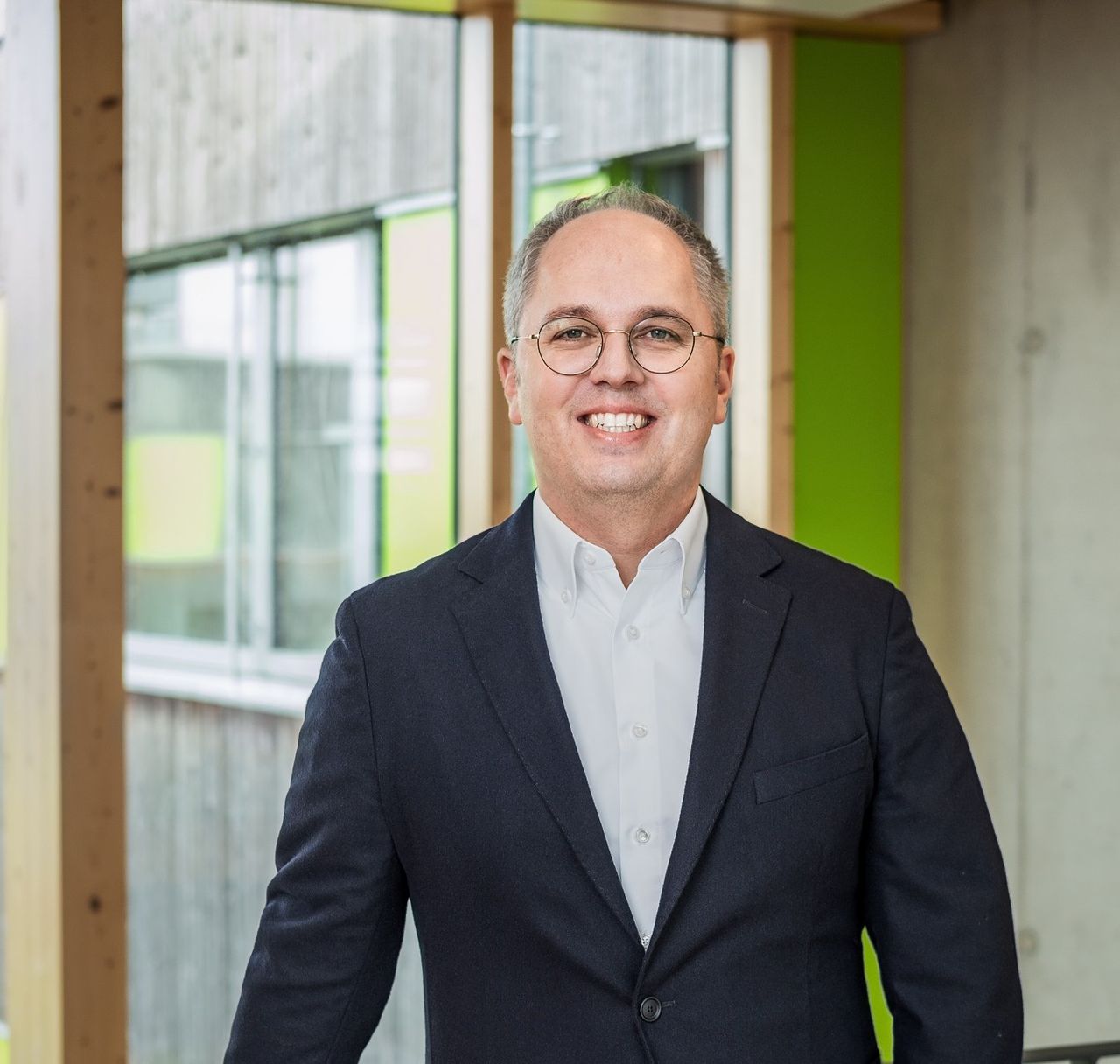 CEO Jens Meier steht an Geländer vor Fenstern und lächelt in die Kamera