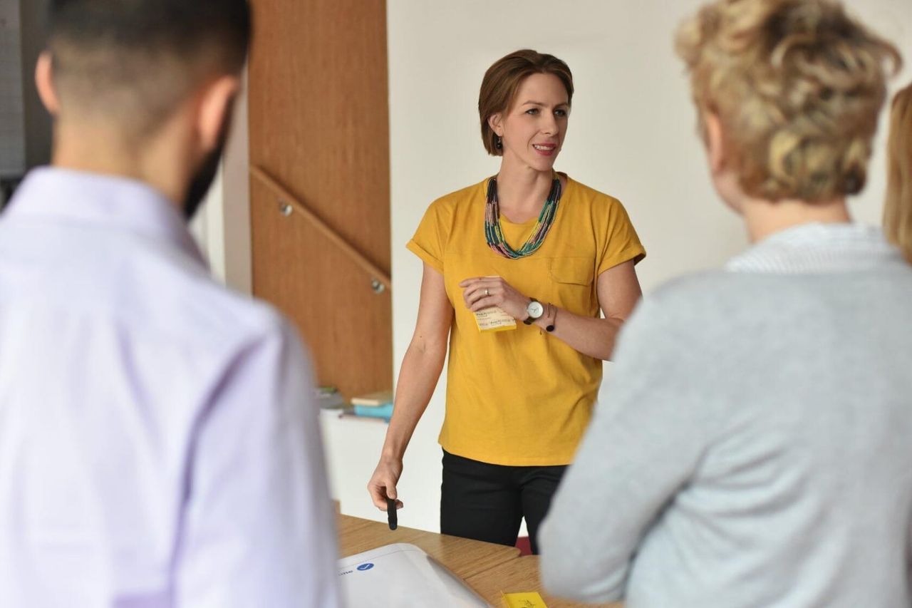 A woman in a yellow shirt stands at a table, engaging with three colleagues in a discussion.