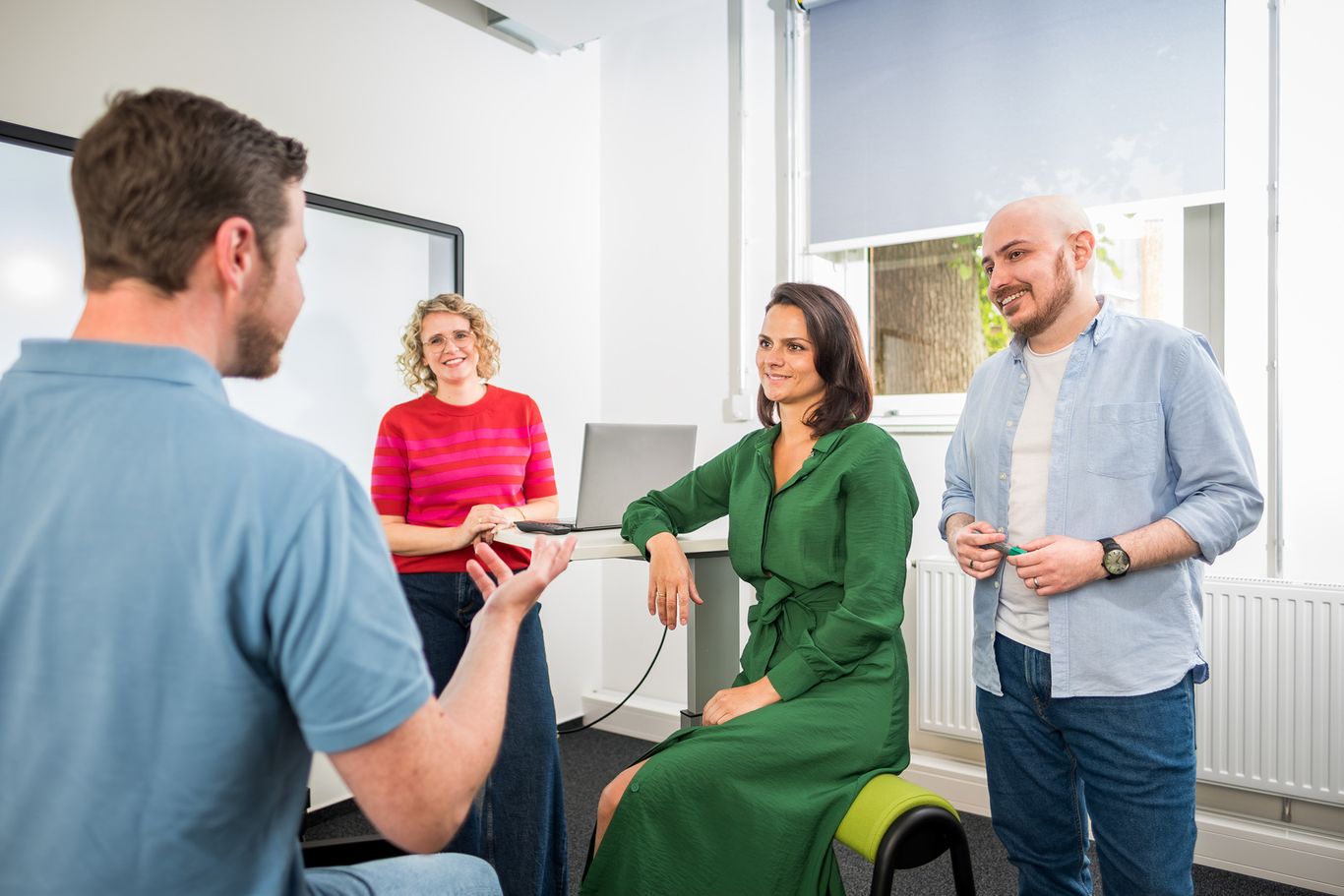 Four people engaged in a casual meeting in a bright office, with one person speaking and others listening attentively.