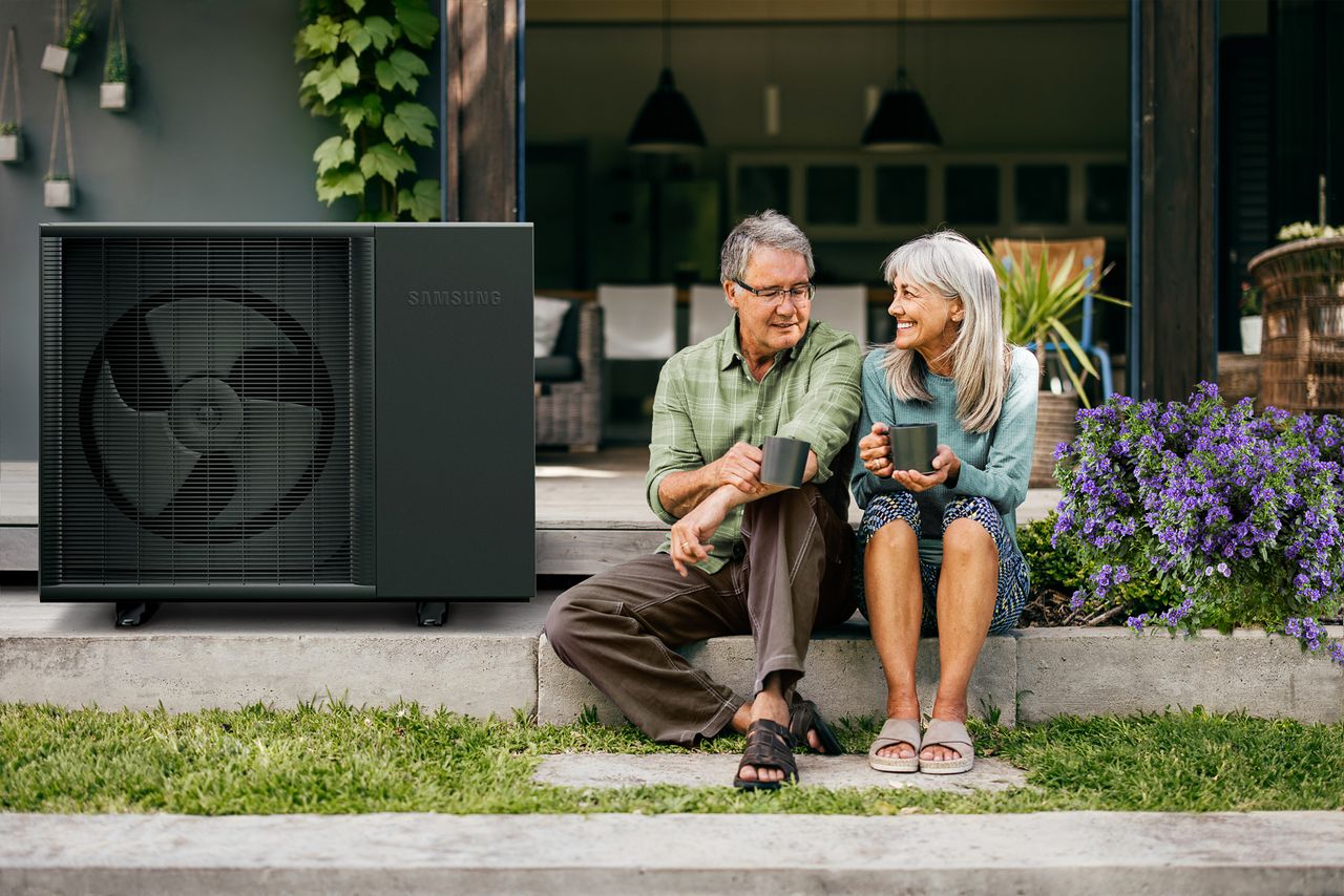 Paar sitzt mit Tassen vor einem Haus, neben ihnen steht eine Wärmepumpe