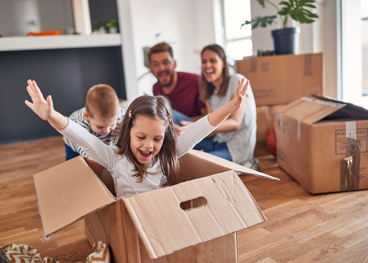 Ein fröhliches Mädchen spielt in einem Umzugskarton, umgeben von seiner lächelnden Familie und weiteren Kartons in einem hellen Raum.
