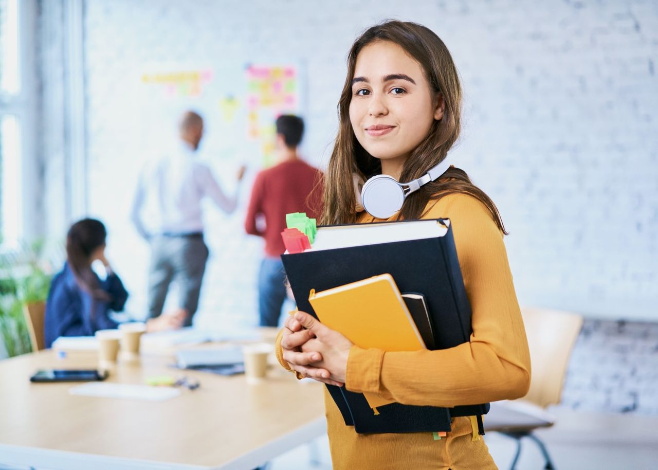 Junge Studentin mit Büchern im Arm schaut in Kamera. Im Hintergrund sind andere junge Menschen am Arbeiten.