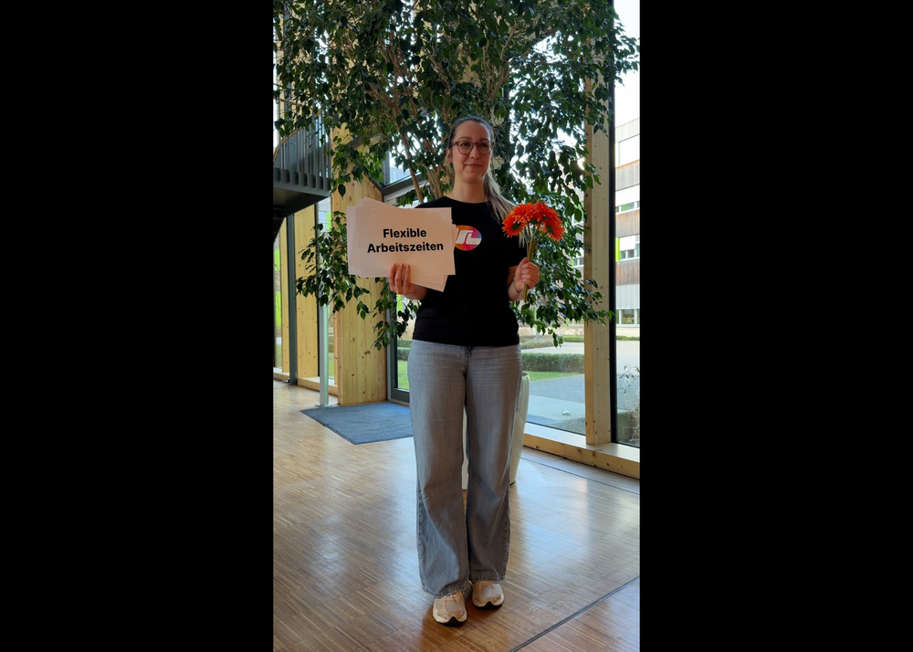 Person holding a sign that reads "Flexible Arbeitszeiten" and a bouquet of orange flowers, standing in a modern wooden building interior.