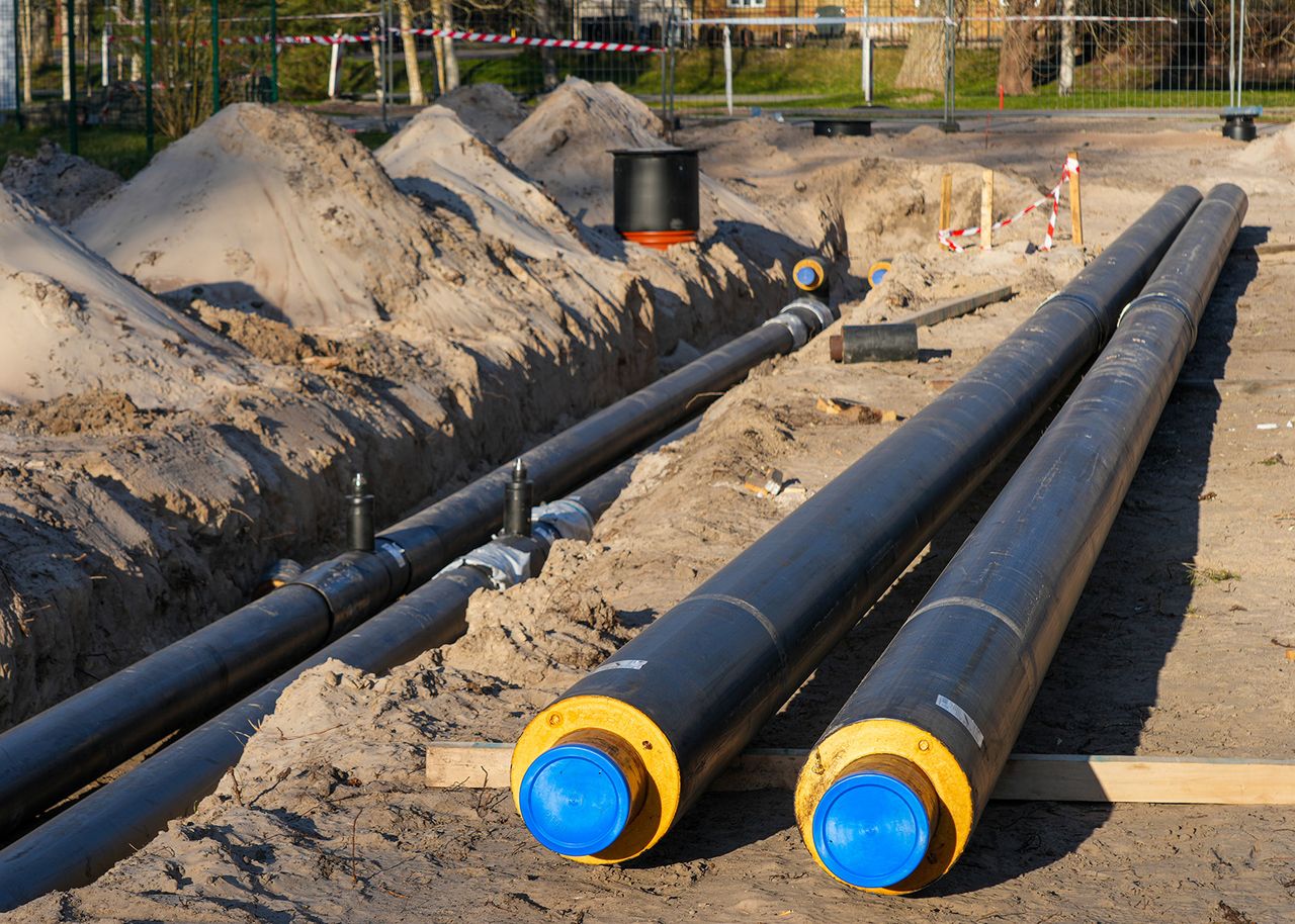 Auf einer Baustelle liegen große schwarze Fernwärme-Rohre mit blauen Kappen in einem Graben, umgeben von Aushub.