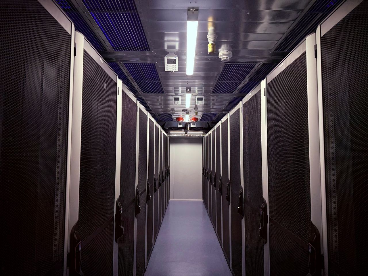 Ein Gang zwischen Reihen hoher Server-Racks in einem schwach beleuchteten Rechenzentrum, mit Deckenbeleuchtung und sichtbarer Deckeninfrastruktur.