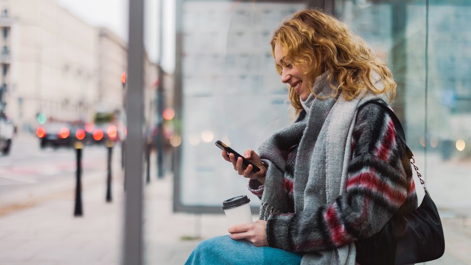 Frau an Bushaltestelle lächelt auf ihr Handy