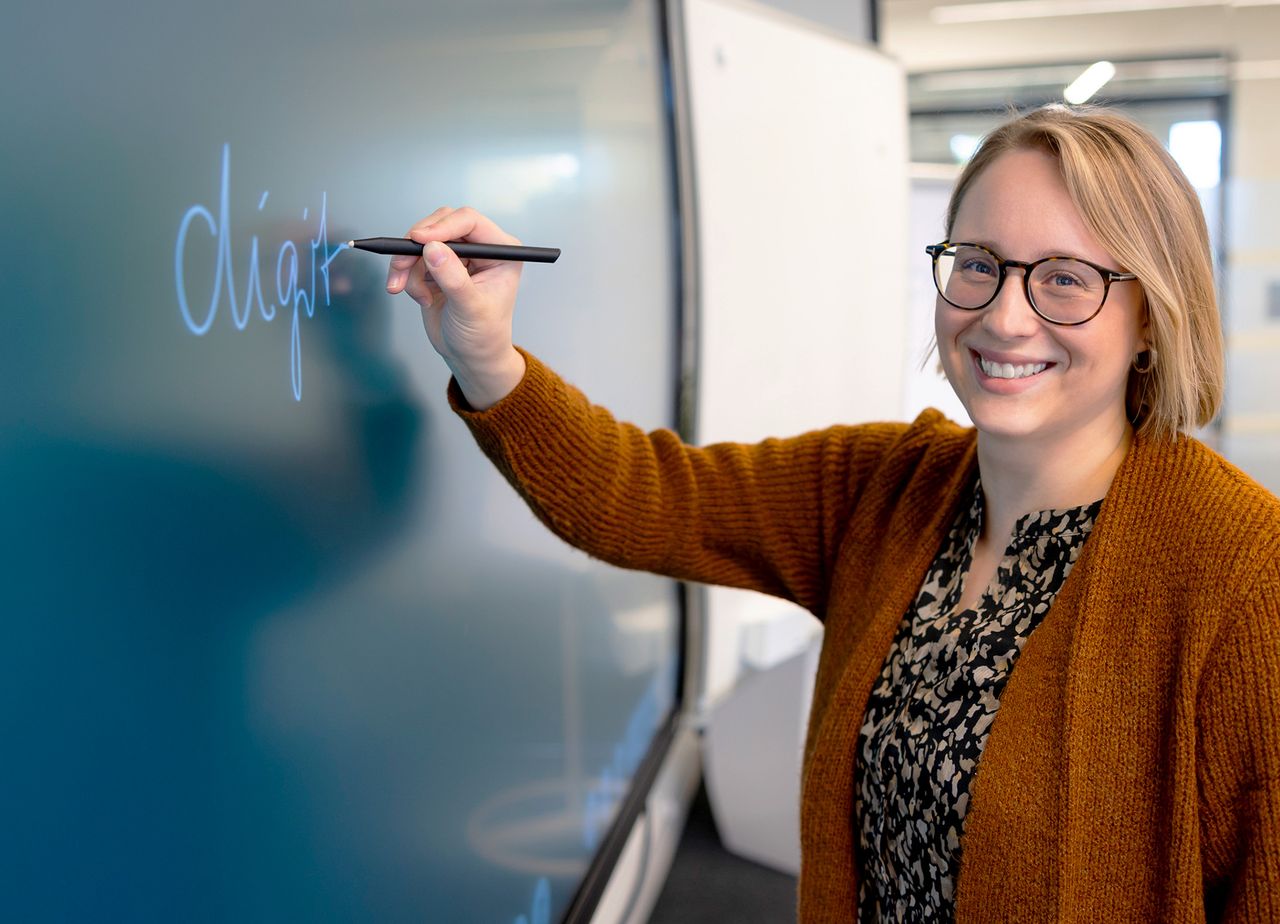 A woman in glasses and a brown sweater writes "digit" on an interactive whiteboard with a stylus, smiling at the camera.