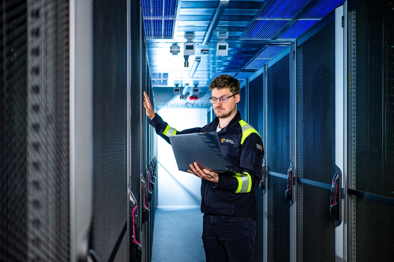 Ein Techniker überprüft Server in einem Gang eines Rechenzentrums; er hält einen Laptop in der Hand und trägt eine dunkle Jacke mit reflektierenden Streifen.
