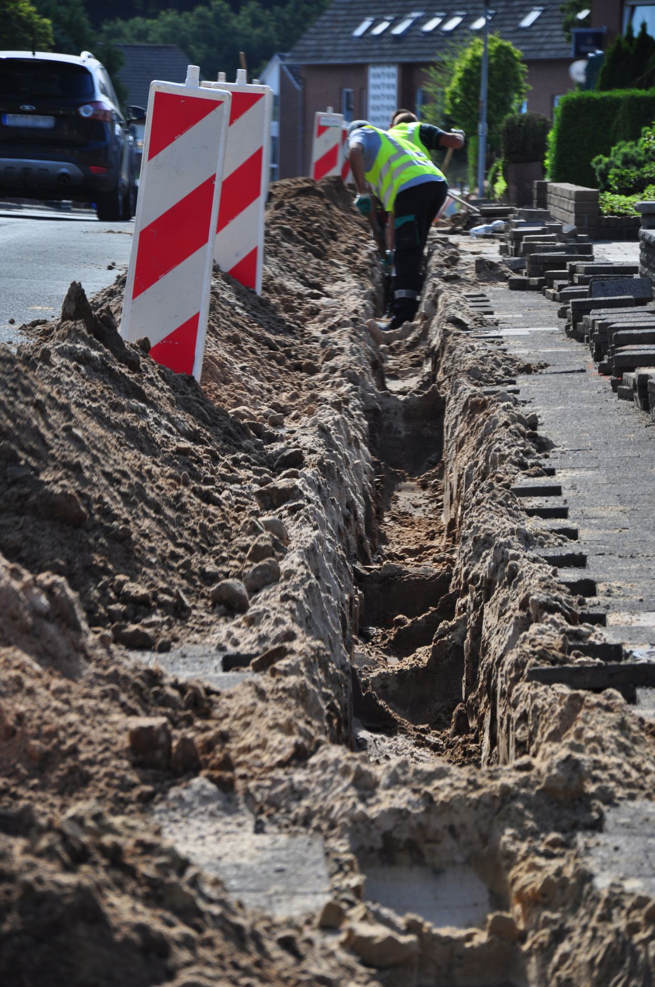 Construction worker digging a trench along a roadside, with traffic barriers and sand piles lining the excavation.