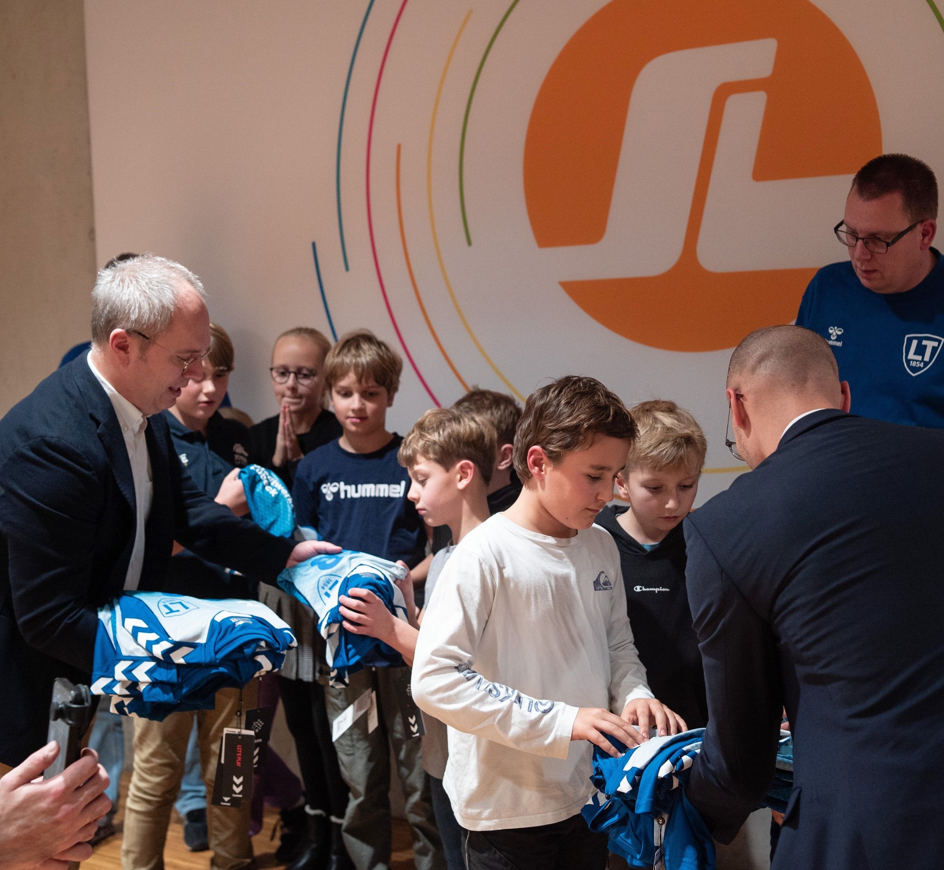 A group of children receive blue sports jerseys from two men in suits, standing in front of a wall with a circular logo.