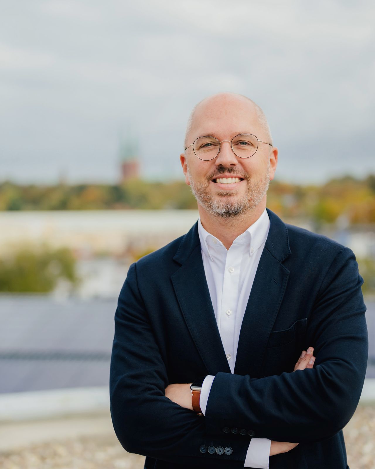 A man in a black blazer and glasses stands outdoors with arms crossed, smiling, with a blurred cityscape in the background.