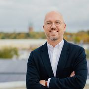A man in a black blazer and glasses stands outdoors with arms crossed, smiling, with a blurred cityscape in the background.
