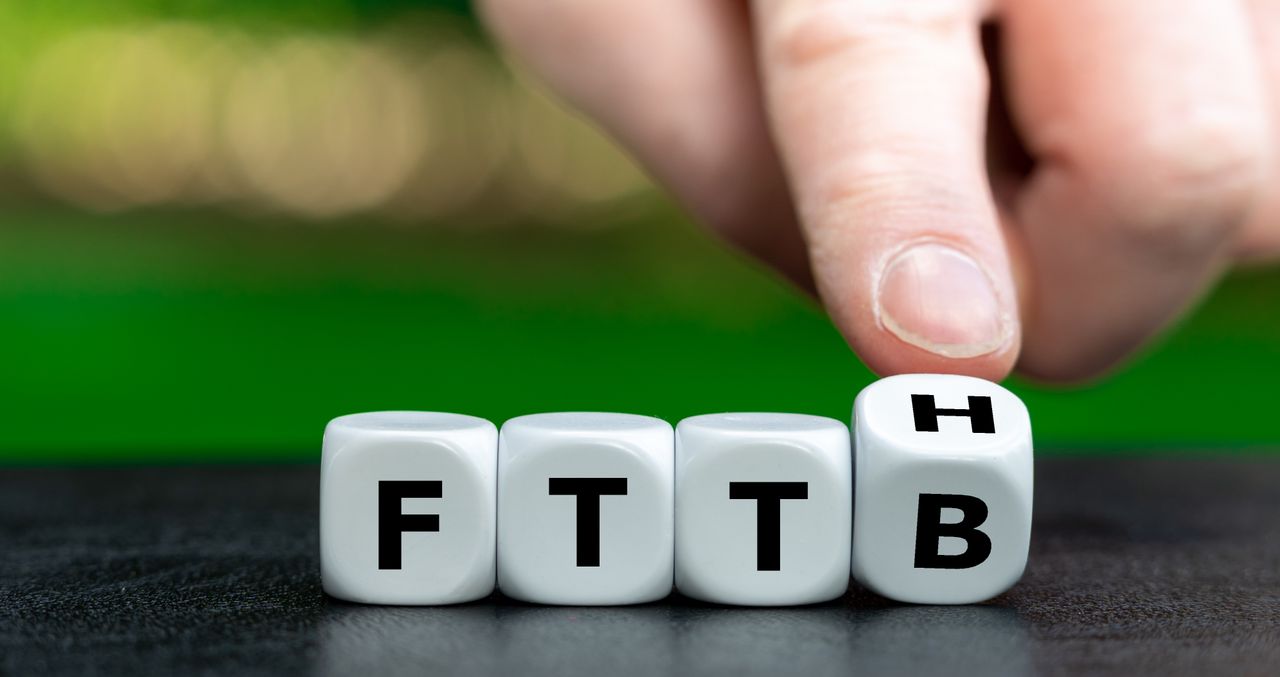 Fingers turning a die from "FTTB" to "FTTH" on a table, with a blurred green background.