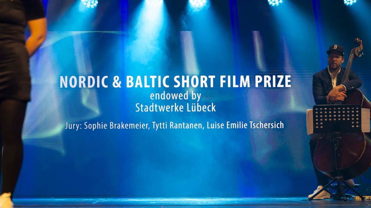 Stage with "Nordic & Baltic Short Film Prize" text, a musician with a double bass, and partial view of a person walking.