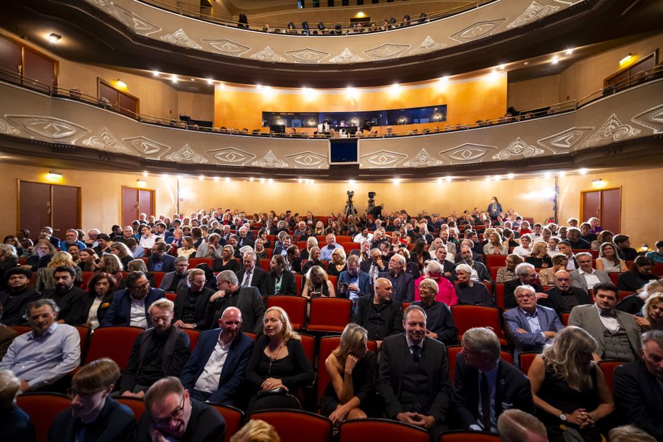 Audience seated in a grand theater with ornate balconies, awaiting a performance. The atmosphere is lively and anticipatory.