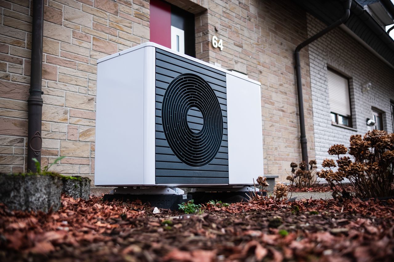 A modern white heat pump unit with a black circular vent is installed outside a brick building, surrounded by fallen leaves.