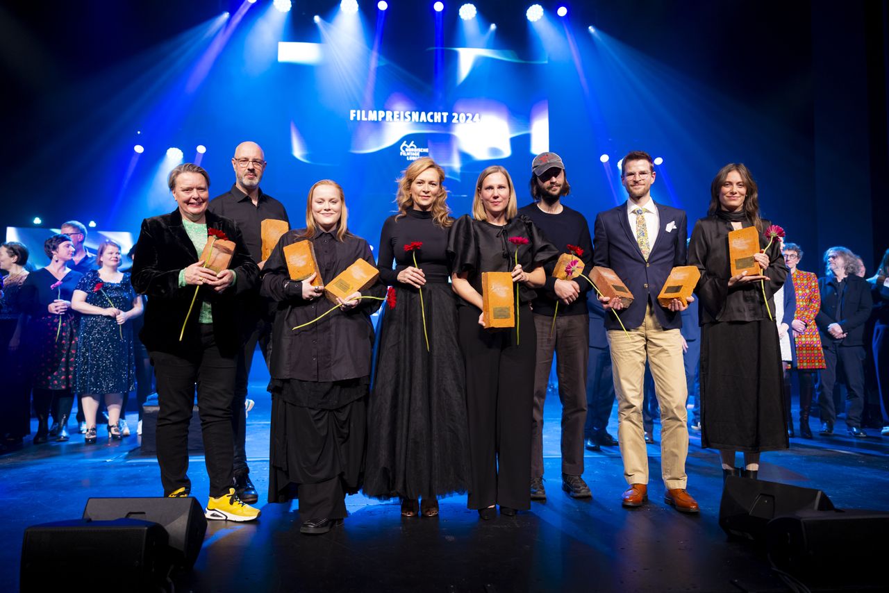 A group of people standing on stage holding awards, with a lit backdrop displaying "Filmpreisnacht 2024."