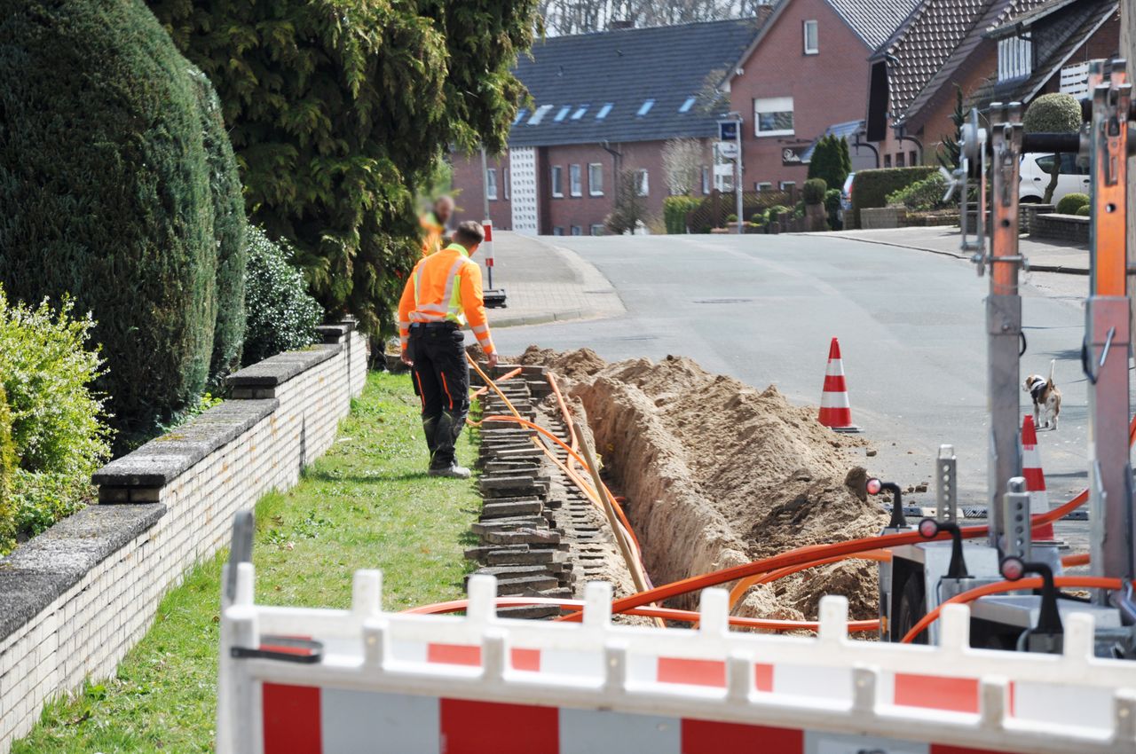 Worker in high-visibility clothing installs cables in a trench along a residential street, with traffic cones and barriers marking the area.
