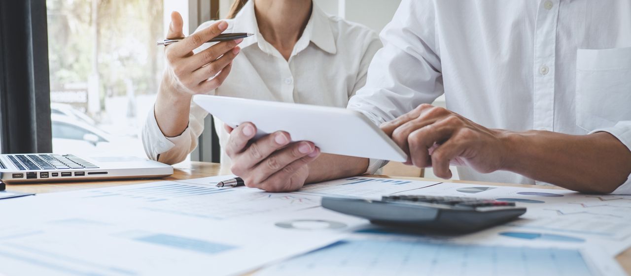 Two people in white shirts discussing data on a tablet, with documents, a calculator, and a laptop on the table.