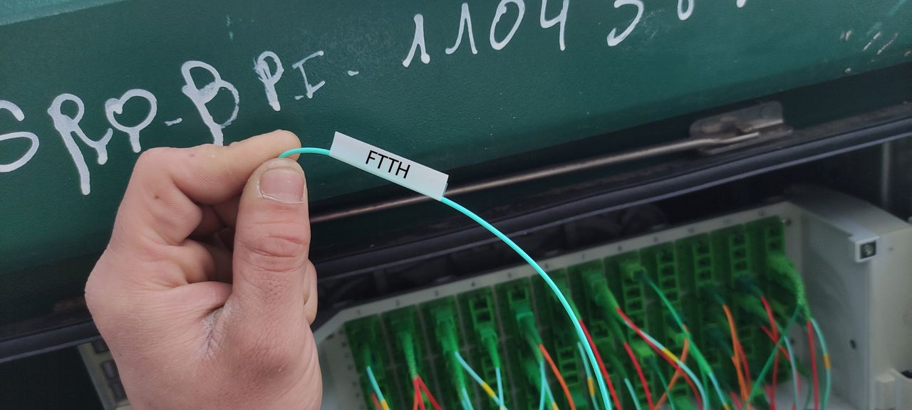 Hand holding a fiber optic cable labeled "FTTH" near a panel with multiple green connectors and handwritten white text on a green surface.