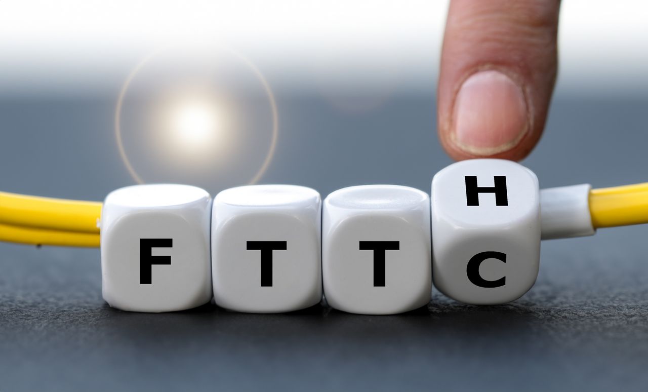 A hand flips a white dice from "FTTC" to "FTTH" against a blurred background, with a yellow cable beneath the dice.