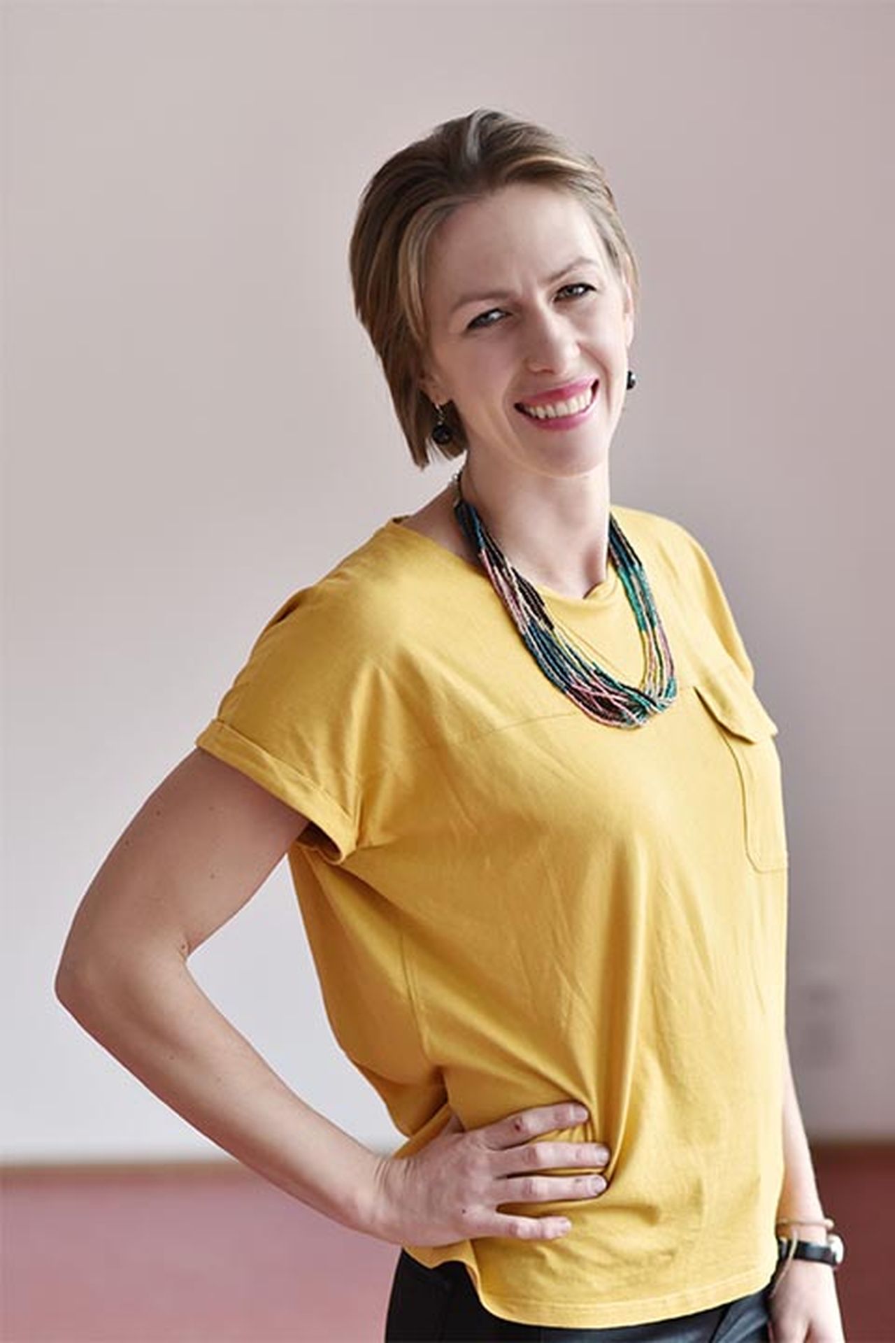 Lila Sax in a yellow shirt and colorful necklace, standing with one hand on hip against a plain background.