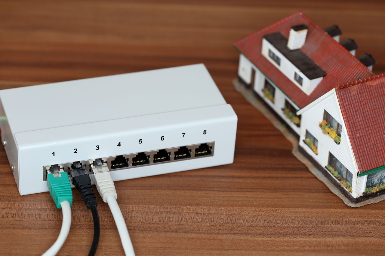 A network switch with cables plugged in, next to a small model house on a wooden surface.