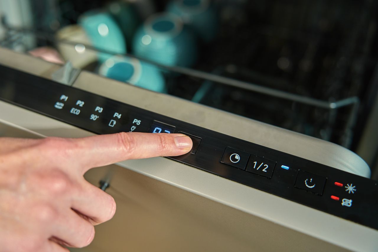 A person presses a button on a dishwasher control panel, with blue dishes visible inside the open dishwasher.