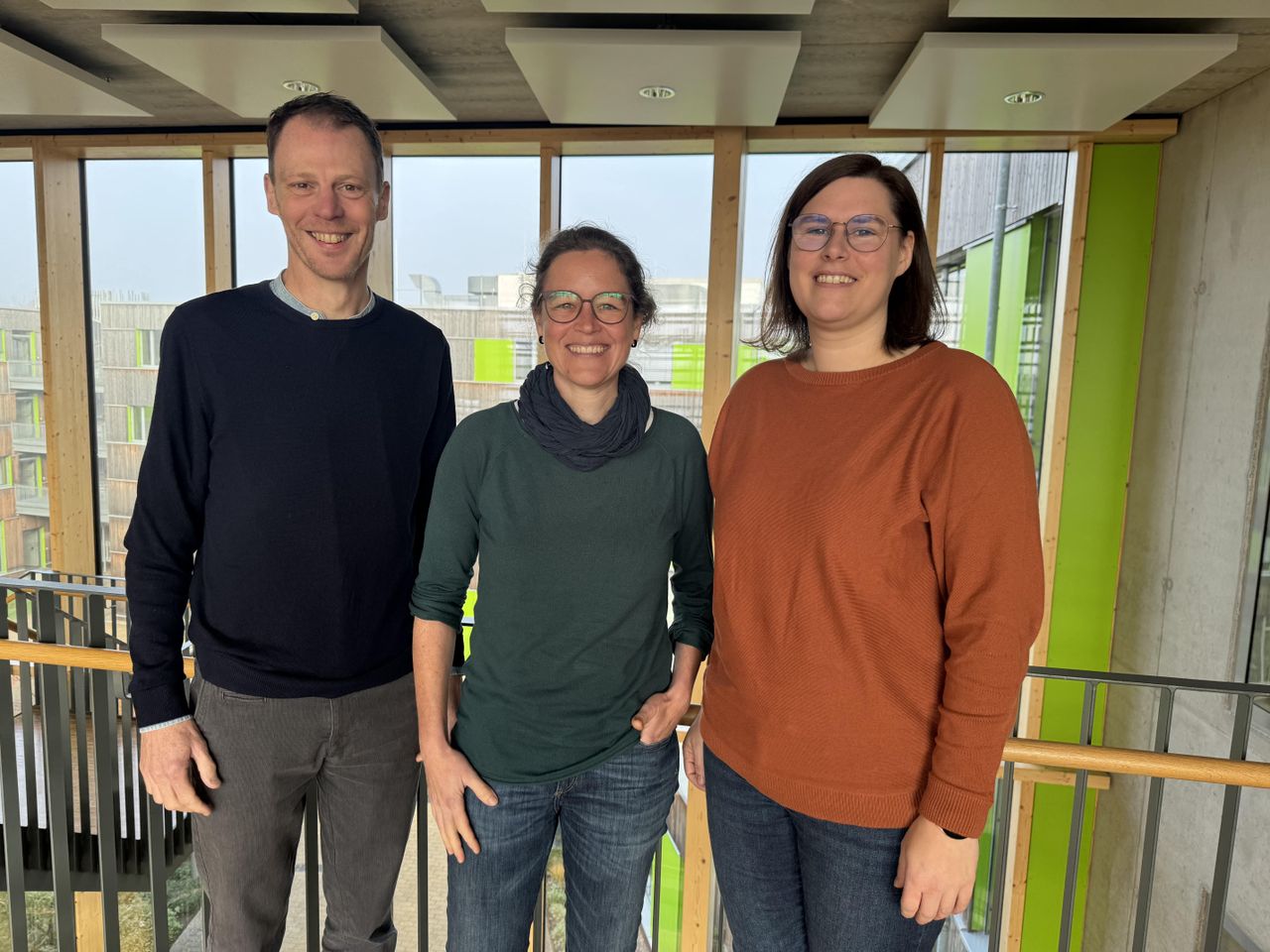 Three people standing indoors, smiling at the camera. They are casually dressed, with a wooden railing and large windows in the background.