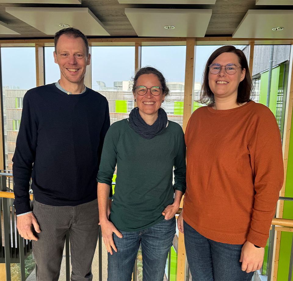 Three people standing indoors, smiling, with a modern building interior and large windows in the background.