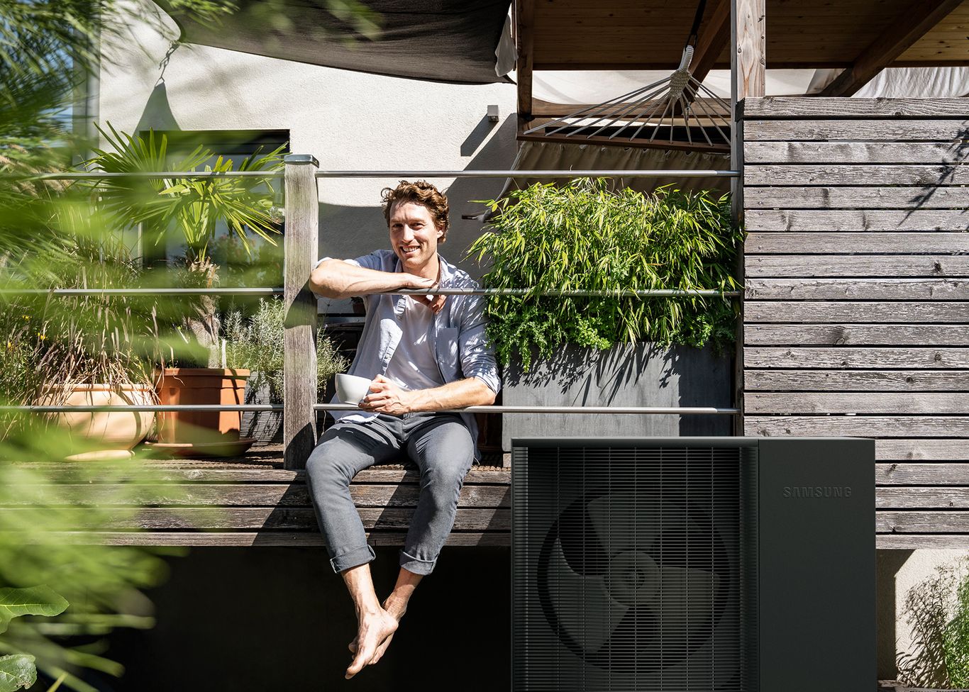 Ein Mann sitzt auf einer Holzveranda, hält eine Tasse in der Hand, ist von Pflanzen umgeben, daneben ist eine Wärmepumpe zu sehen
