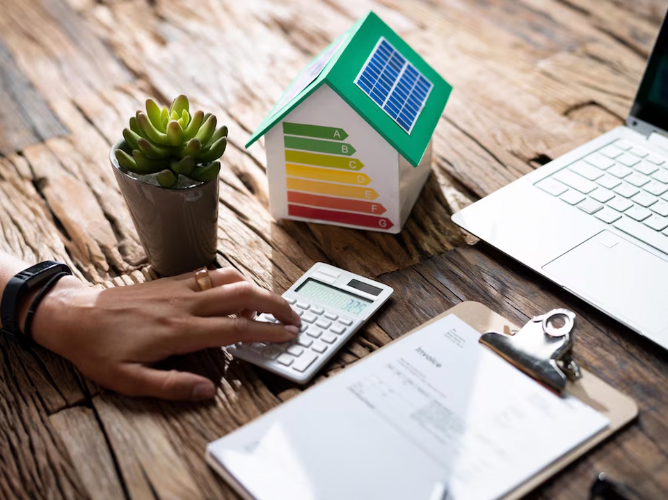 Person using a calculator at a wooden desk with a clipboard, laptop, potted plant, and a model house displaying energy efficiency ratings.