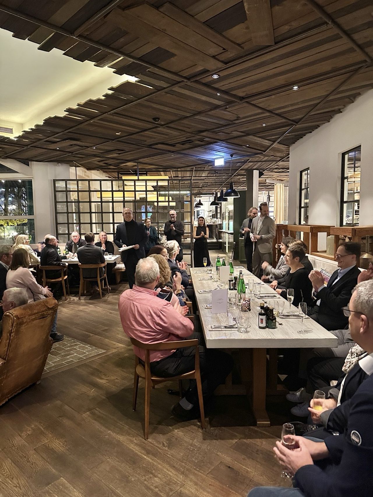 A group of people seated around a long dining table in a restaurant, listening to a speaker. The room has a rustic wooden ceiling design.