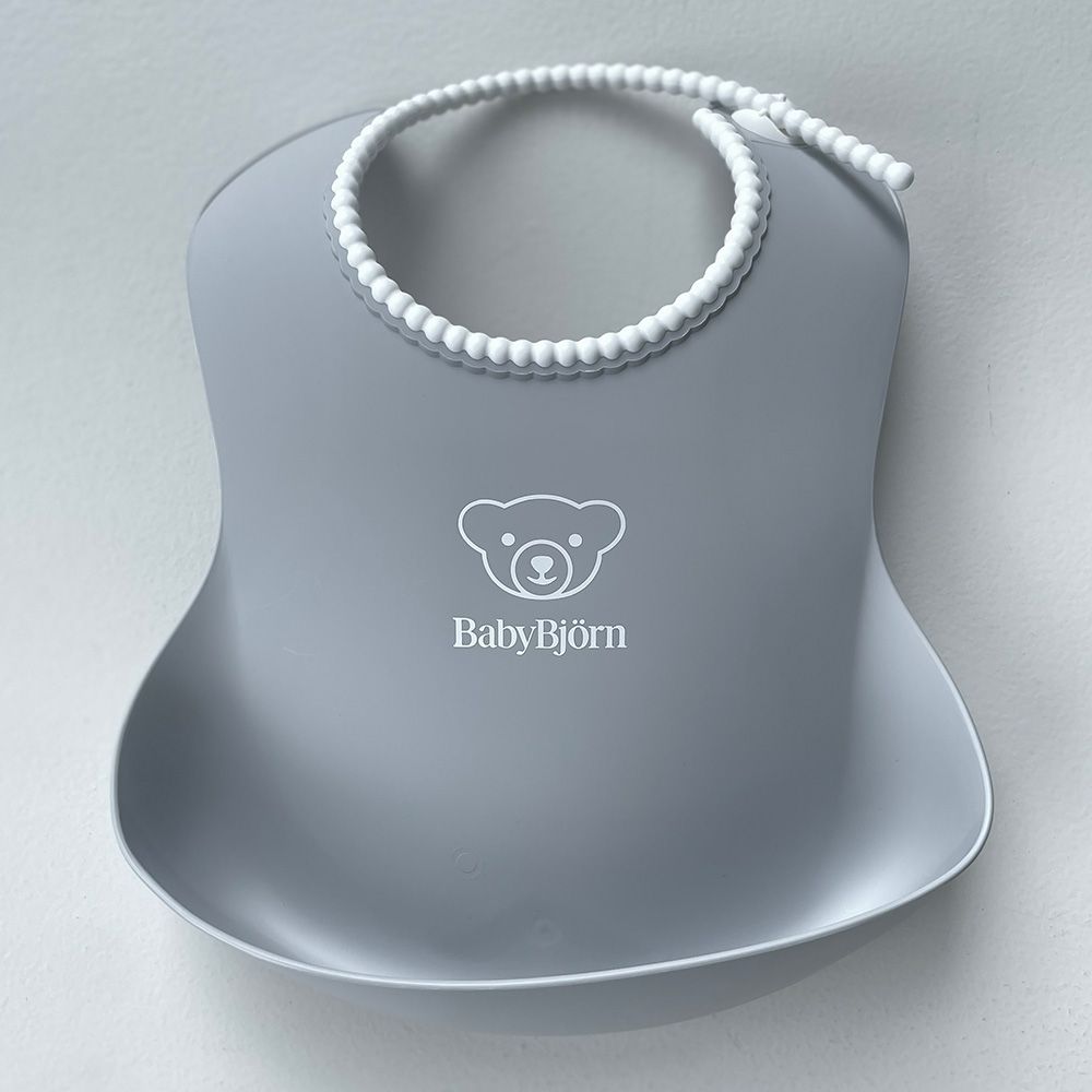 Gray BabyBjörn bib with a bear logo and a beaded adjustable strap, designed to catch food spills.