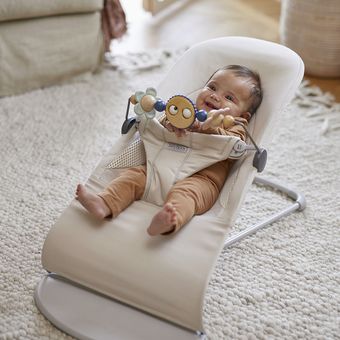 Baby sitting in a beige rocker on a rug, smiling while holding a colorful toy with a sun face, indoors.