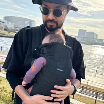 Man wearing sunglasses and a hat holds a baby in a black carrier by a riverside, with modern buildings in the background.