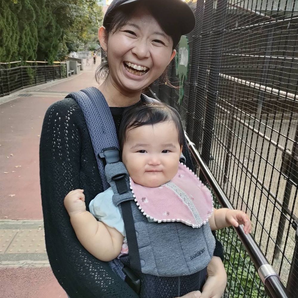 A smiling woman wearing a cap holds a baby in a carrier on a sidewalk, both looking happy. The baby wears a pink bib.