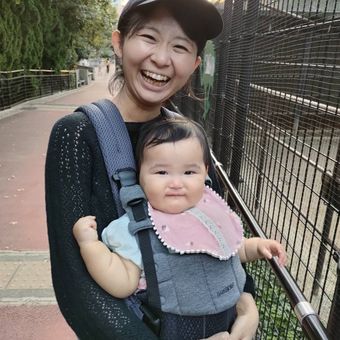 A smiling woman wearing a cap holds a baby in a carrier on a sidewalk, both looking happy. The baby wears a pink bib.