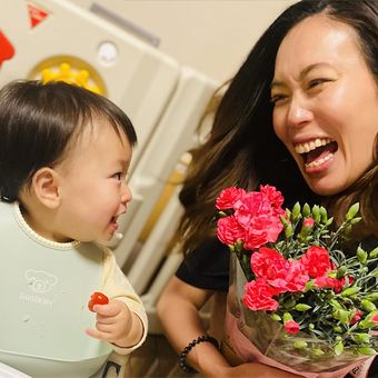A woman joyfully holds pink flowers while smiling at a baby holding a small toy, both sharing a happy moment indoors.