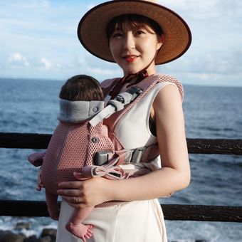 Woman in a sun hat holds a baby in a pink carrier by the ocean, with a wooden railing and blue sky in the background.
