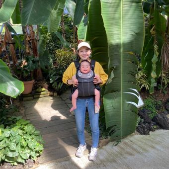 A woman in a yellow sweater and cap carries a baby in a front carrier, standing on a garden path surrounded by large green leaves.