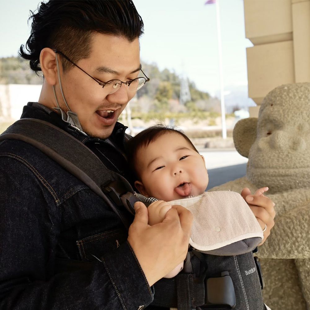 A man smiles at a baby in a carrier who is playfully sticking out their tongue. They stand next to a stone sculpture.