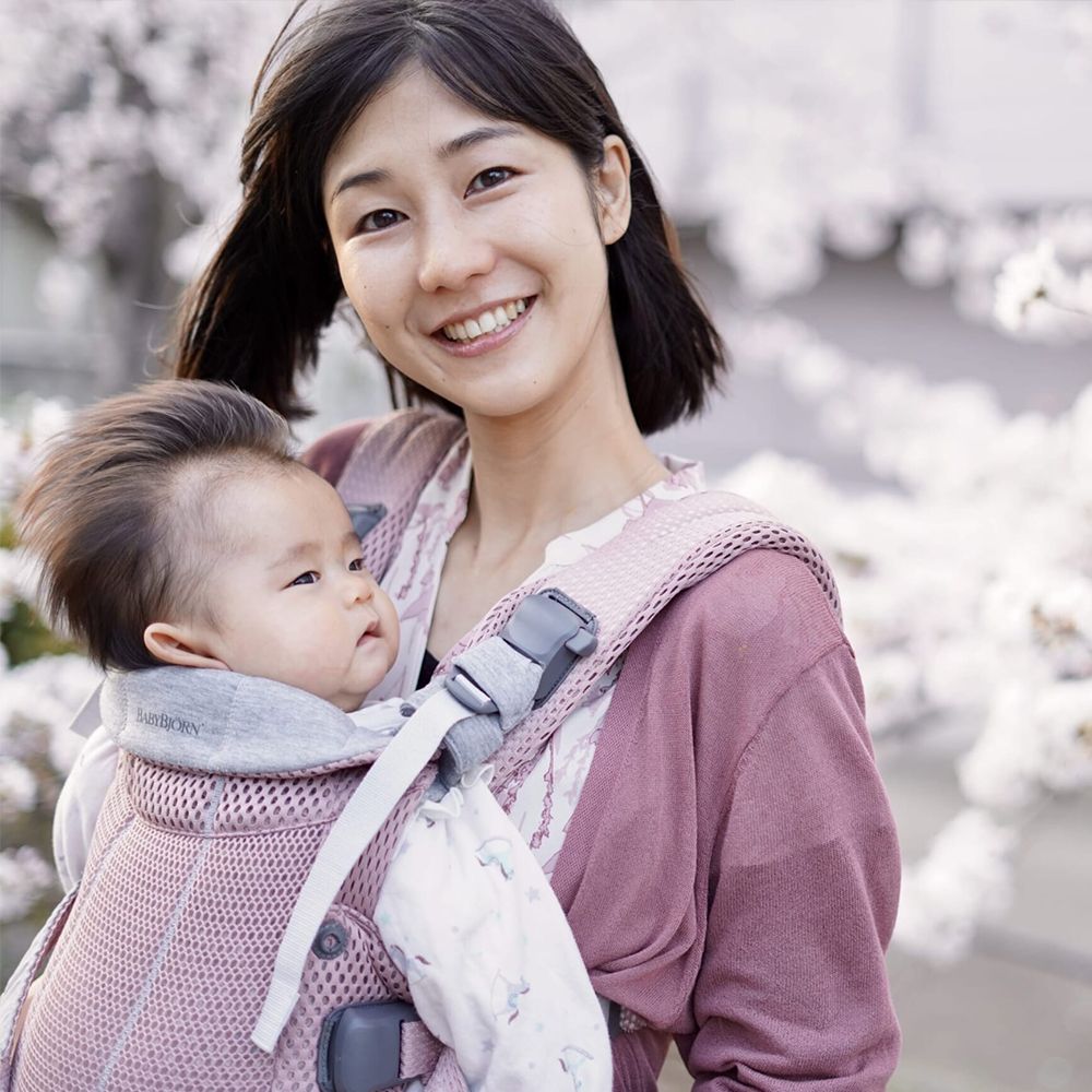Smiling woman carrying a baby in a front-facing carrier, surrounded by blooming cherry blossoms.