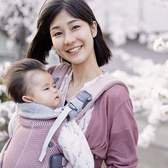 Smiling woman carrying a baby in a front-facing carrier, surrounded by blooming cherry blossoms.