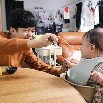 A person smiling and feeding a baby in a high chair with a spoon in a cozy room.